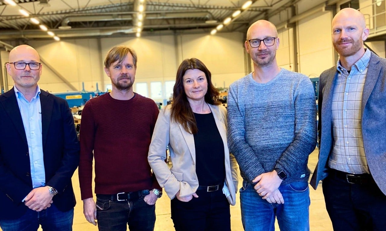 Fem personer står tillsammans i en ljus industrimiljö, klädda i vardags- och kontorskläder. De ler och poserar för kameran.