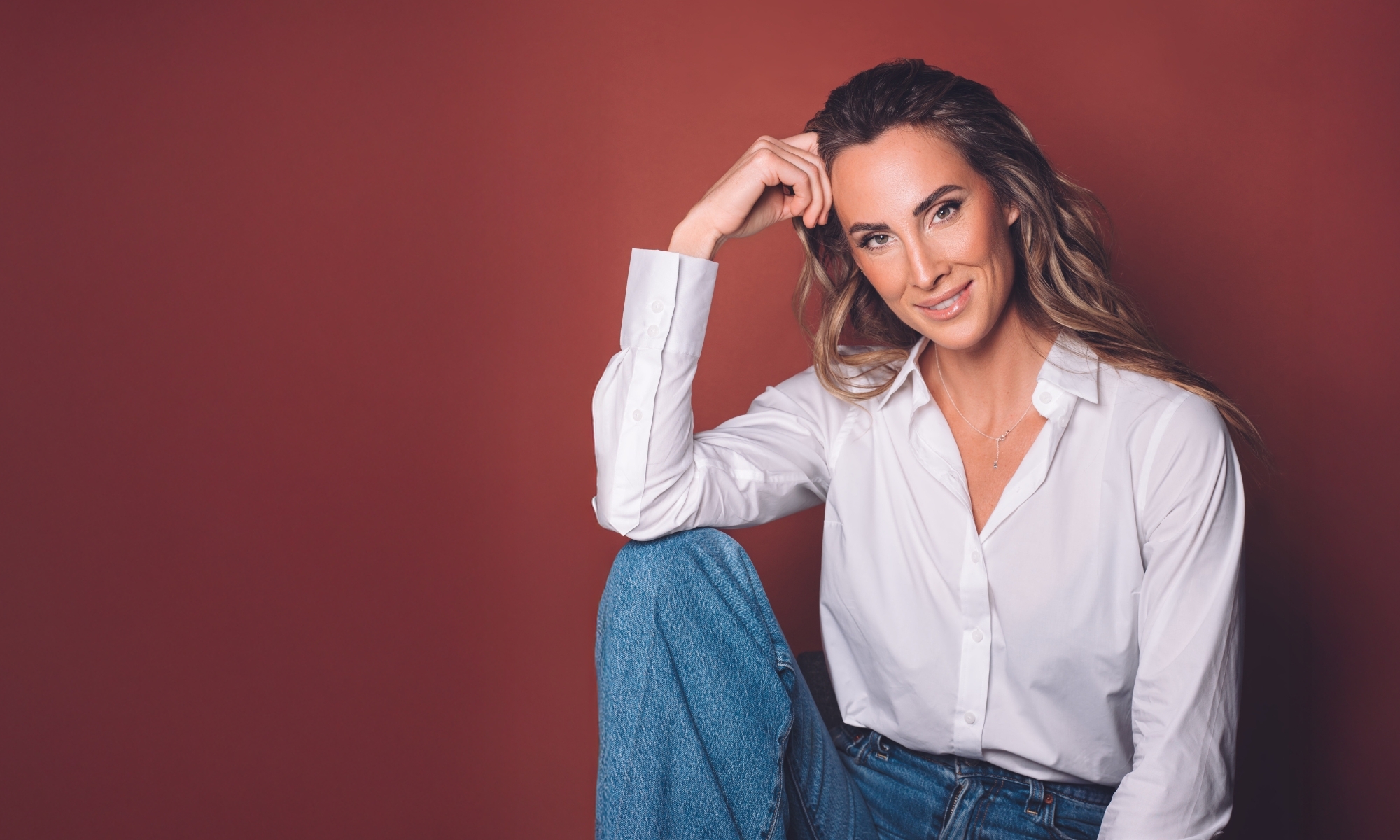 Leende kvinna i vit skjorta och jeans sitter mot röd bakgrund.