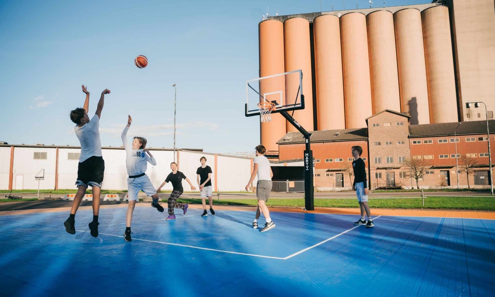 Teens playing basketball on an outdoor court near silos in the evening sun.