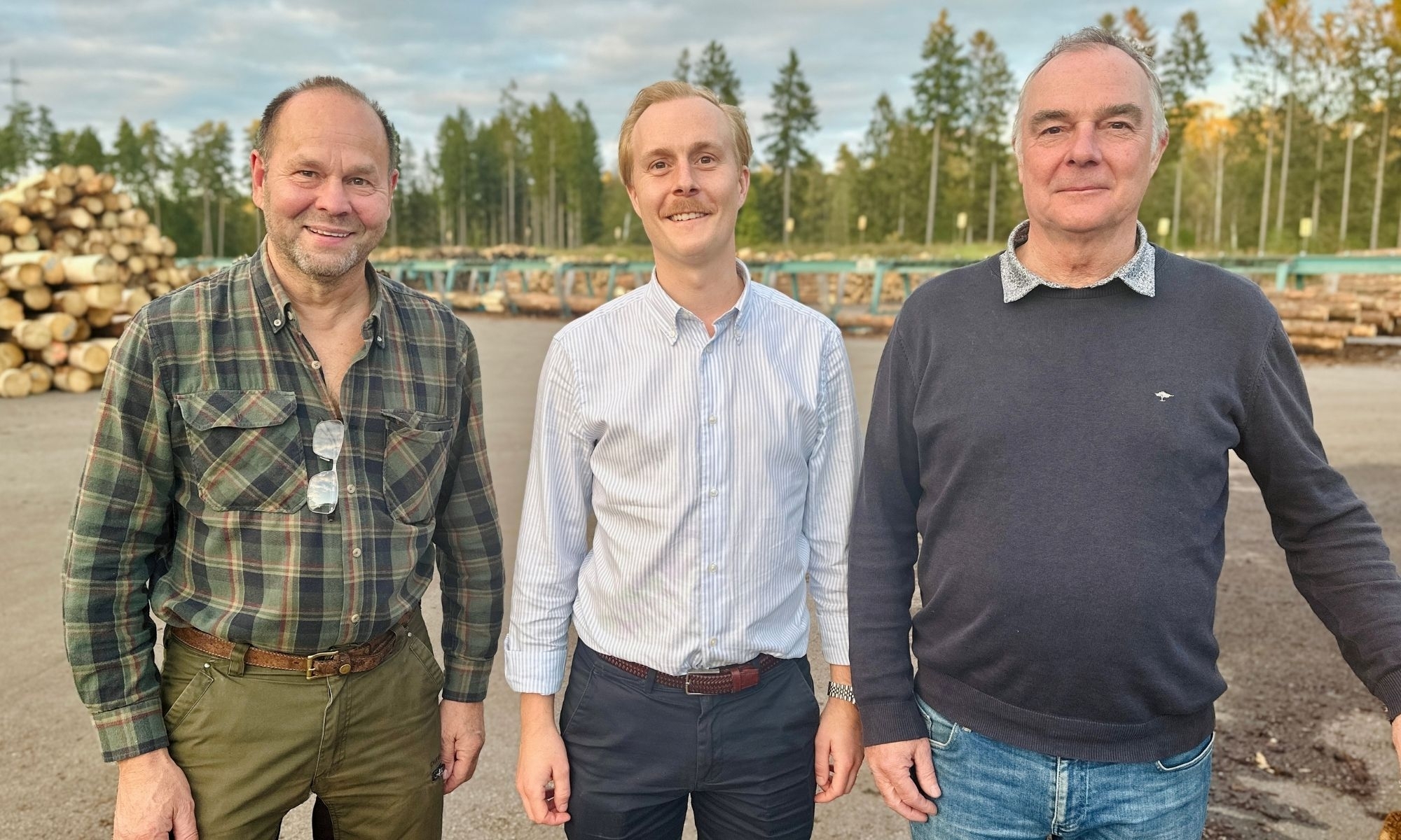 Tre män står framför travar med timmer på ett sågverksområde.