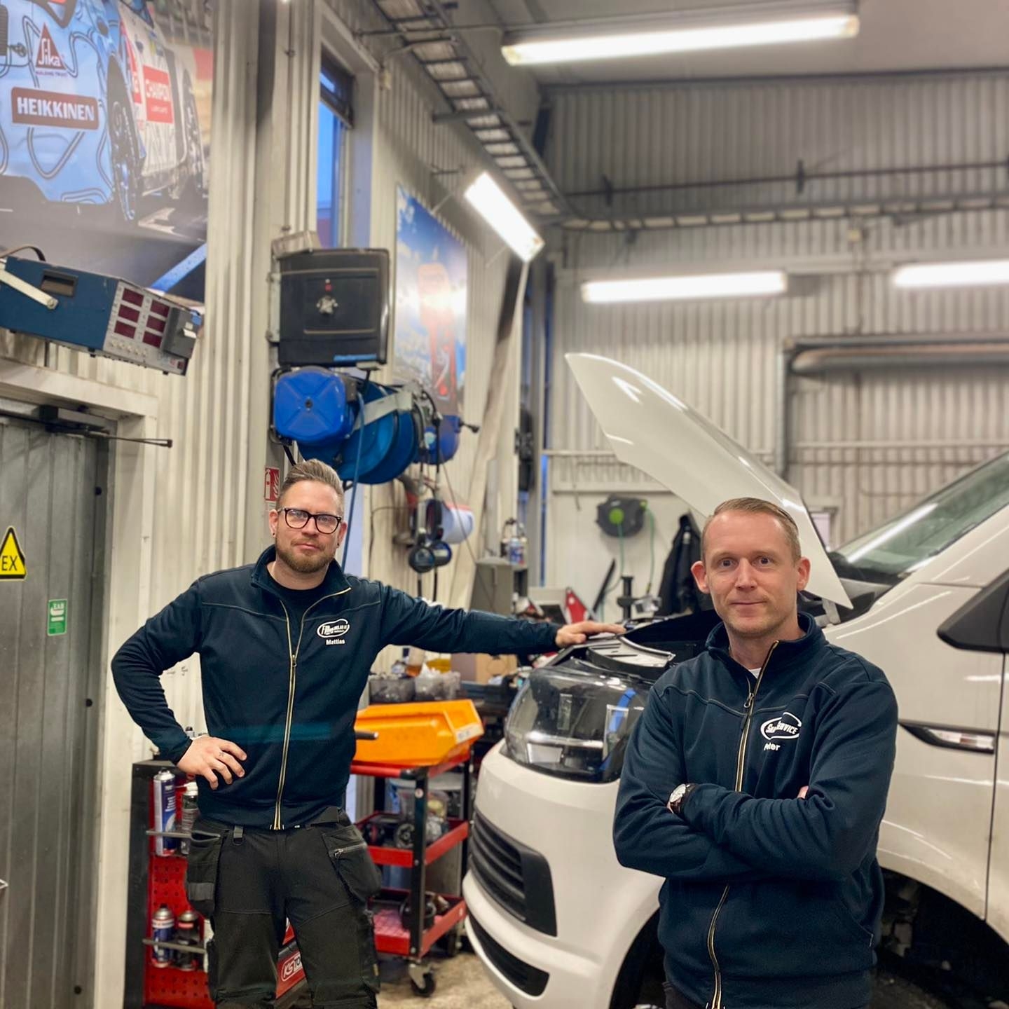 Två män i ett garage står bredvid en vit skåpbil med öppen huv. De har matchande jackor och poserar framför verktyg och utrustning.