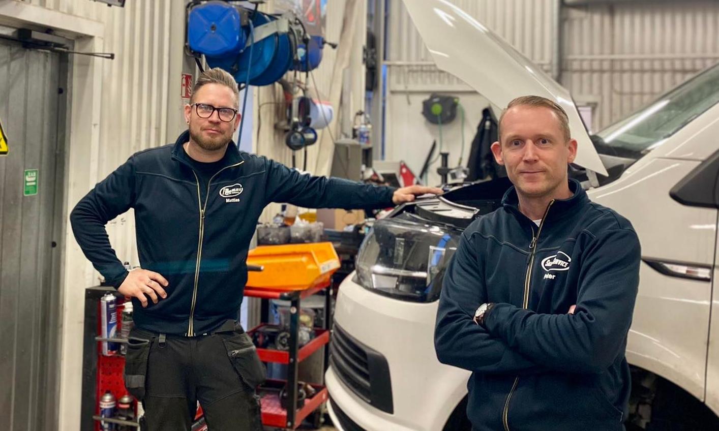 Två män i ett garage står bredvid en vit skåpbil med öppen huv. De har matchande jackor och poserar framför verktyg och utrustning.