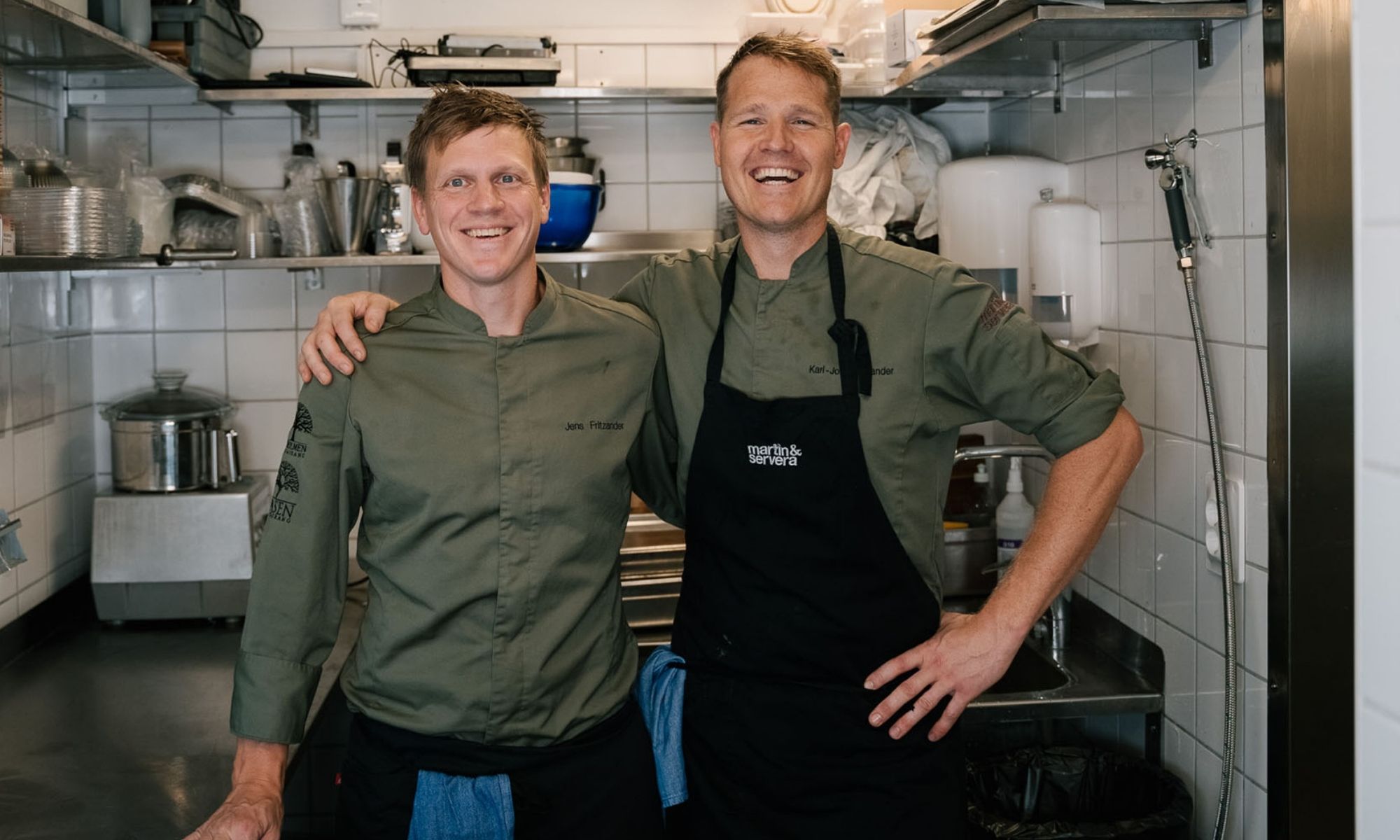 Två kockar står arm i arm och ler i ett restaurangkök, omgivna av rostfria bänkar och köksutrustning.