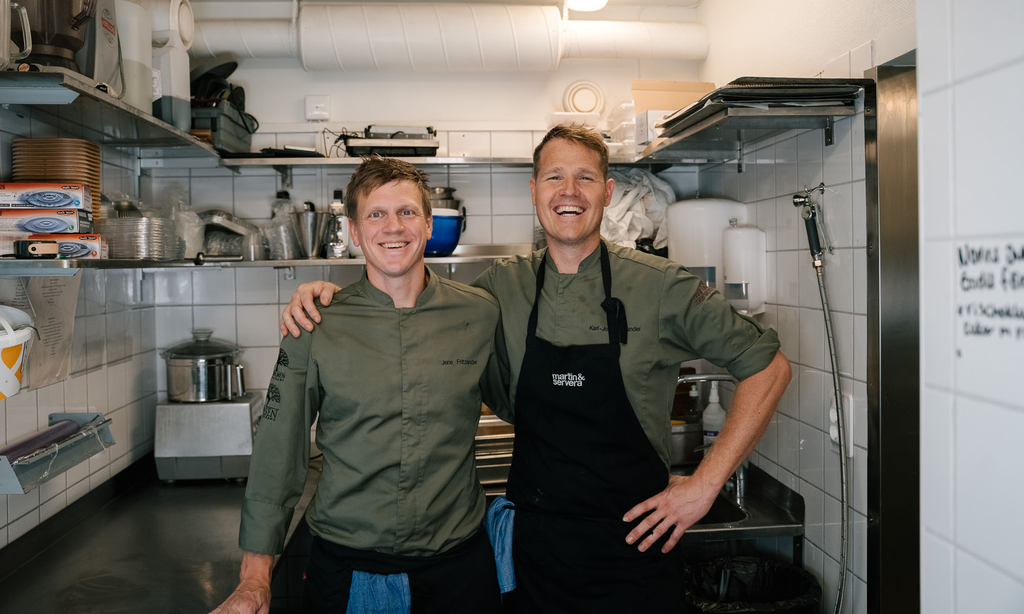 Två kockar står leende i restaurangkök med rostfria bänkar och hyllor.