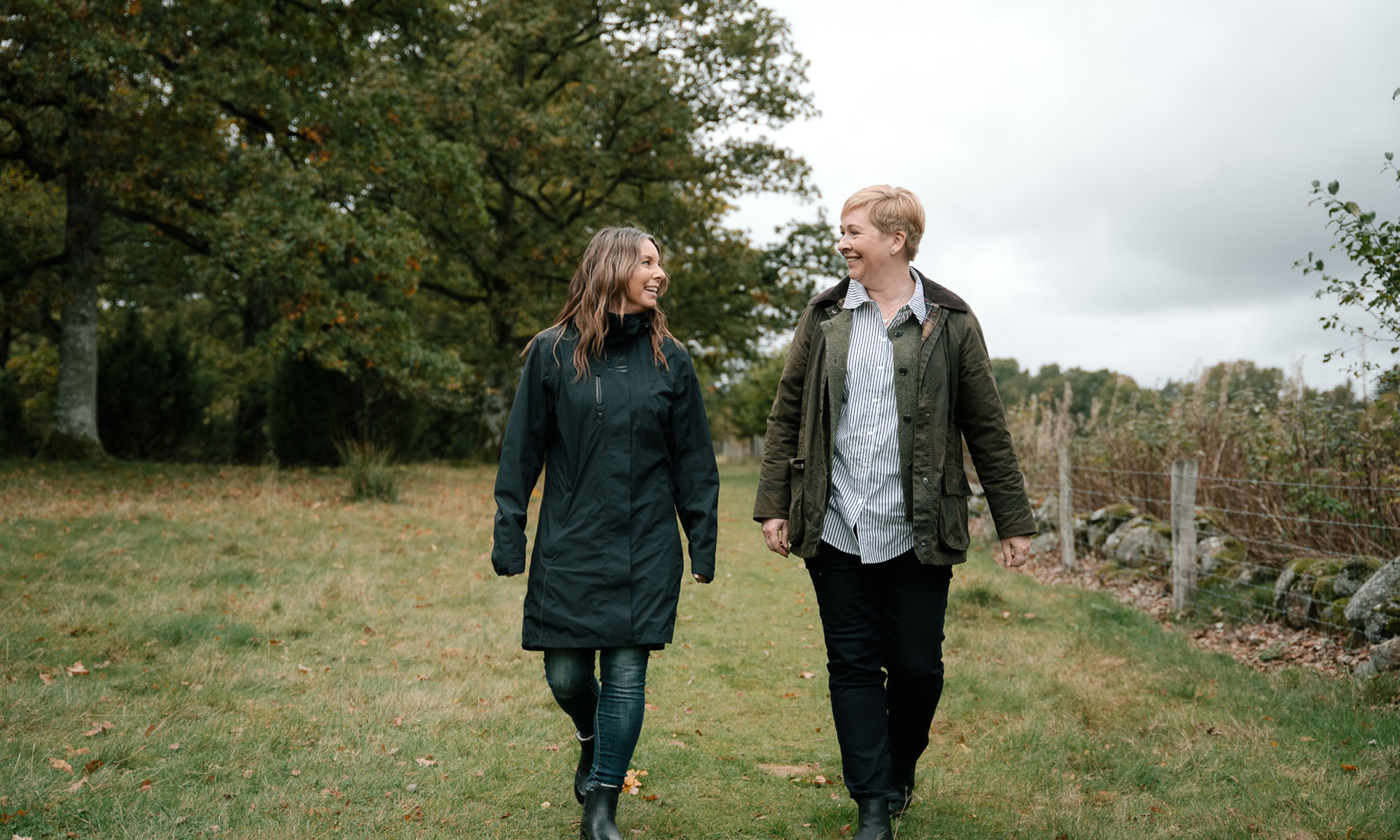Två kvinnor promenerar och samtalar i grönskande landskap.