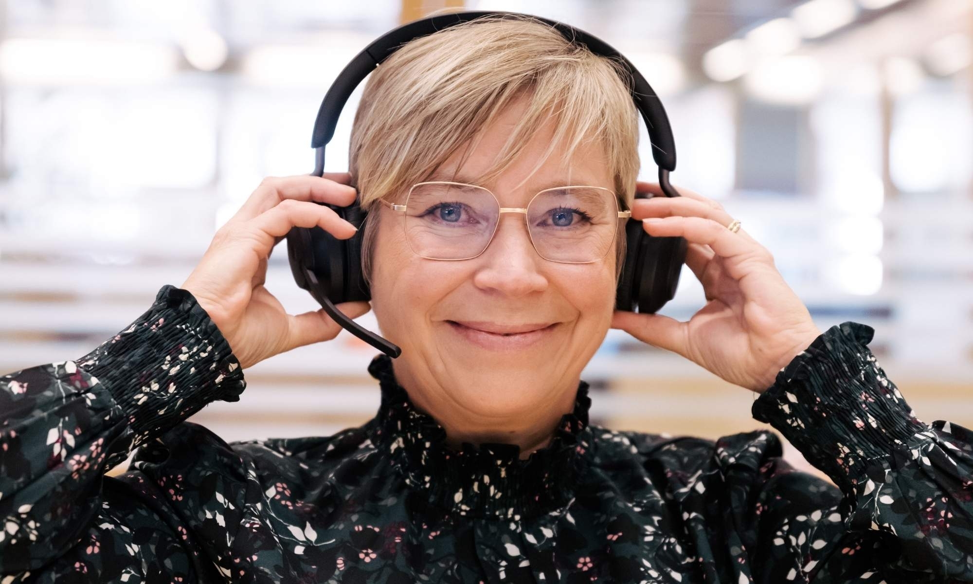 En leende kvinna med kort blont hår och glasögon justerar sitt headset i en ljus kontorsmiljö.