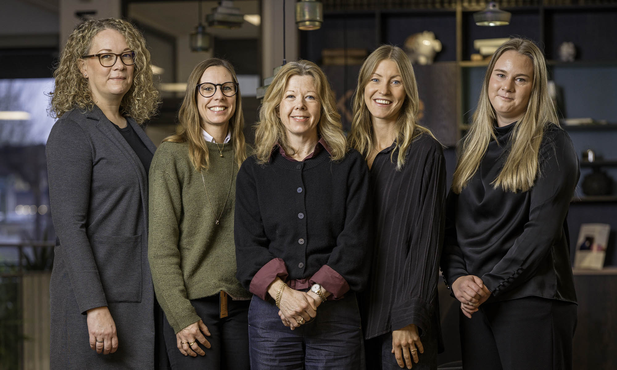 Fem kvinnor står tillsammans i en modern kontorsmiljö och ler mot kameran.