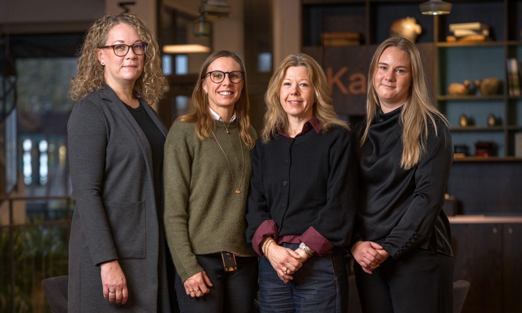 Fyra kvinnor står tillsammans i en varm kontorsmiljö och ser in i kameran.